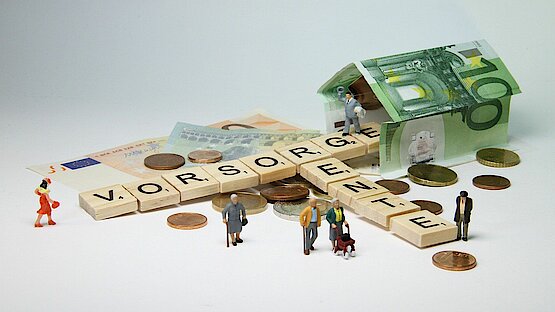 Vorsorge und Rente als Scrabblewörter zusammen gesetzt dazu ein Haus aus 100 Euro scheinen und kleine Figuren ringsrum ein paar Münzen