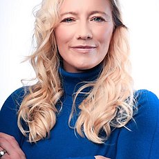 junge Frau mit langen blonden Haaren und blauen Oberteil vor weißer Wand