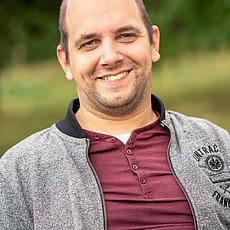 Mann mittleren Alters mit grauer Jacke udn dunkelrotem Oberteil vor grünem Hintergrund